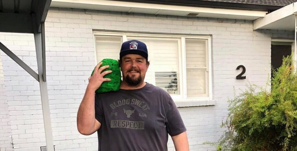 dwayne holding bright green rock heading off for a walk, infront of his house with a big smile